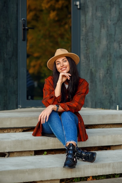 La bella donna in camicia, jeans e cappello rossi si siede sulle scale della sua casa moderna vicino alla foresta.