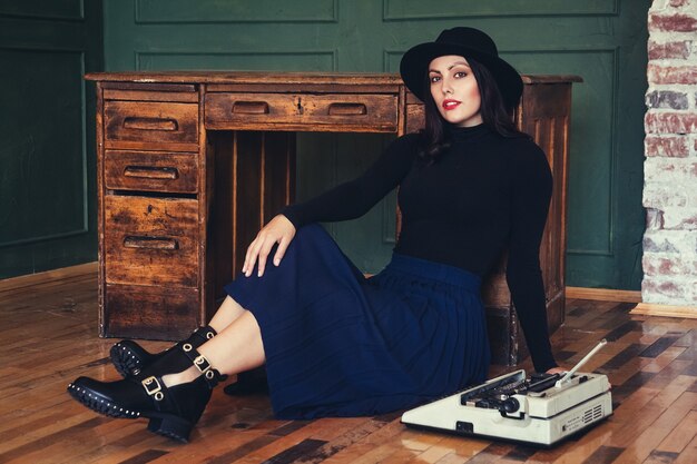 La bella donna in un cappello si siede vicino al tavolo di quercia con la macchina da scrivere vintage.