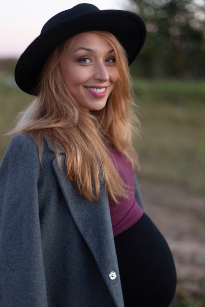 La bella donna incinta nei sorrisi del cappotto e del cappello, esamina la macchina fotografica fuori