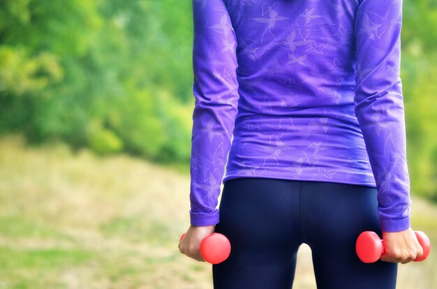 La bella donna jogger in camicia blu con manubri rosa nelle sue mani esegue l'esercizio sportivo sulla natura verde colorata. DOF poco profondo.