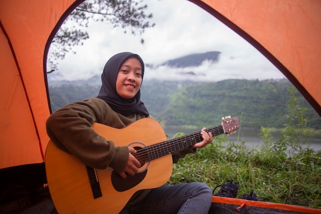 La bella donna musulmana asiatica gioca la chitarra che si siede vicino alla vista del fiume del lago della montagna con lo zaino, tenda al tramonto