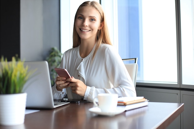 La bella donna sorridente di affari sta sedendosi nell'ufficio e sta esaminando la macchina fotografica.