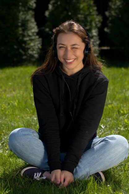 La bella donna sorridente felice in cuffie strizza l'occhio seduto sull'erba all'aperto