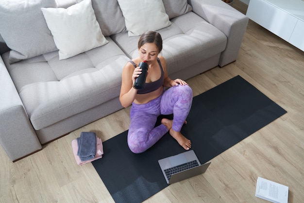 La bella donna sportiva atletica che si siede sulla stuoia di yoga dopo alcuni esercizi beve il frullato proteico o