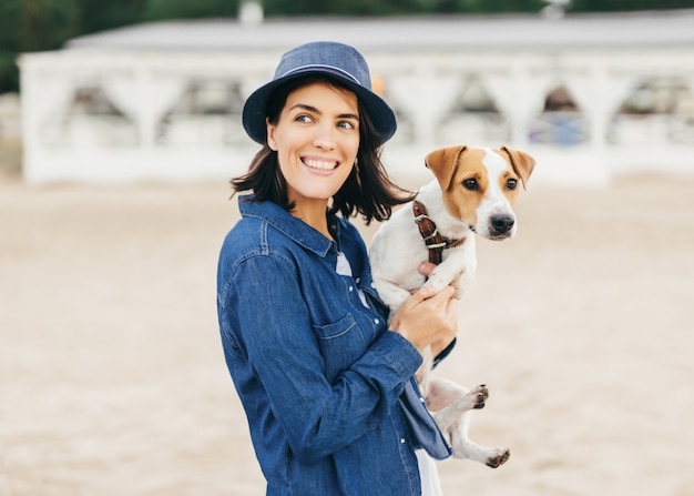 La bella femmina positiva in cappello alla moda e camicia di jeans tiene il suo animale domestico sulle mani, guarda con un sorriso da parte mentre nota la sua amica, cammina all'aperto. Persone, animali, stile di vita e concetto di relax