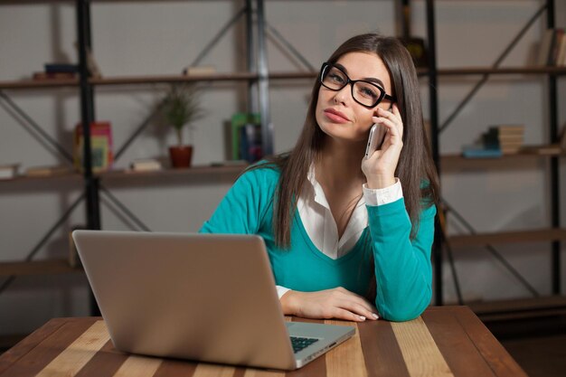 La bella freelance sta lavorando con qualcosa e si siede al tavolo di legno con laptop e telefono