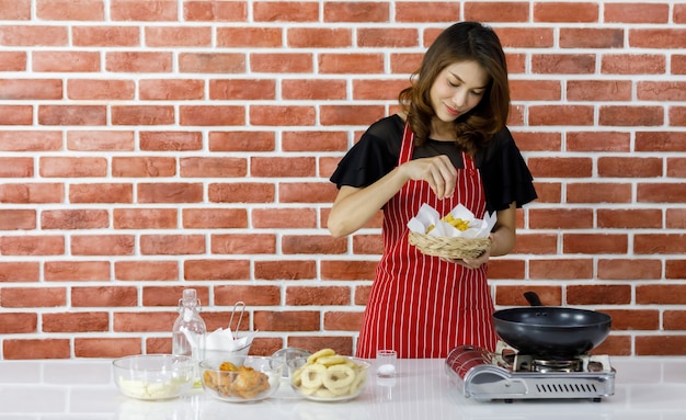 La bella giovane casalinga asiatica in grembiule rosso si diverte a cucinare vicino al muro di mattoni della cucina di casa tenendo sorridente un cesto di vimini di patate fritte e versando felicemente sale per aggiungere un sapore più gustoso