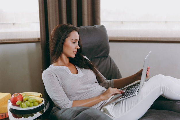 La bella giovane donna a casa seduta su una sedia moderna davanti alla finestra, rilassante nel suo soggiorno e lavorando con il laptop