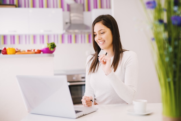 La bella giovane donna allegra sta usando il suo computer portatile mentre teneva la carta di credito e beve il caffè.