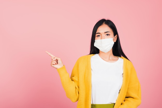 La bella giovane donna asiatica che indossa la protezione della maschera medica previene l'infezione da coronavirus, COVID-19 sta puntando il dito verso lo spazio, girato in studio isolato su sfondo rosa, concetto di assistenza sanitaria medica