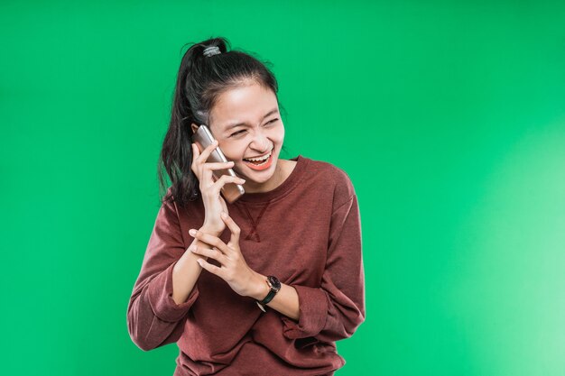 La bella giovane donna asiatica sta parlando al telefono con la risata di espressione felicemente isolata contro i precedenti verdi