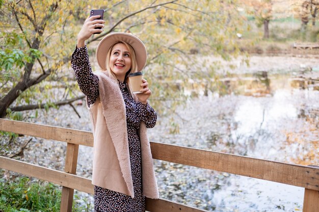 La bella giovane donna beve il caffè prende selfie sul telefono in natura nel parco di autunno in fall