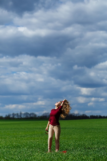 La bella giovane donna bionda sta su erba verde in un parco. Cielo azzurro con nuvole. La ragazza sorride e si gode una buona giornata.