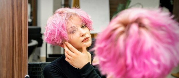 La bella giovane donna caucasica con un nuovo taglio di capelli rosa corto guardando il suo riflesso nello specchio controllando l'acconciatura in un salone di parrucchiere