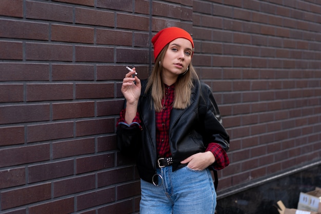 La bella giovane donna caucasica elegantemente vestita fuma la sigaretta sulla strada. Cattiva abitudine al fumo Dipendenza da nicotina Stile di vita malsano. Camminata moderna del bomber e dei jeans della donna all'aperto