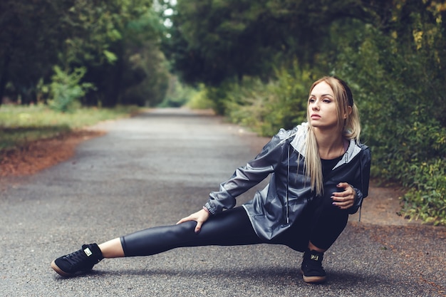 La bella giovane donna che fa l'allungamento si esercita in un parco della città al tempo piovoso.