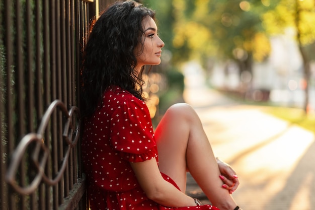 La bella giovane donna con capelli ricci in un vestito d'annata rosso si siede vicino al recinto nel parco al tramonto