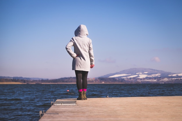 La bella giovane donna con il cappotto grigio sta levandosi in piedi su una passerella e sta godendo del periodo invernale di vista