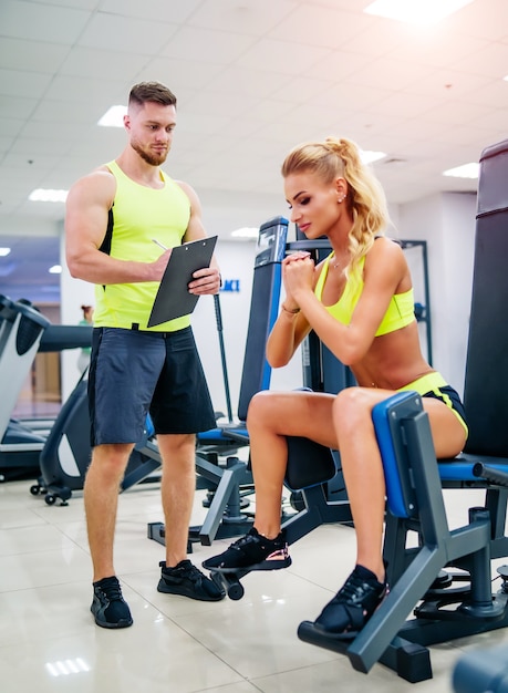 La bella giovane donna con il suo personal trainer in palestra discute i suoi progressi su un blocco per appunti tenuto dall'uomo