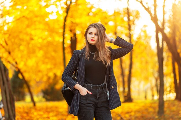 La bella giovane donna con le labbra rosse in un vestito elegante di modo con un blazer, un maglione e uno zaino cammina nel parco con fogliame giallo-arancio variopinto al tramonto