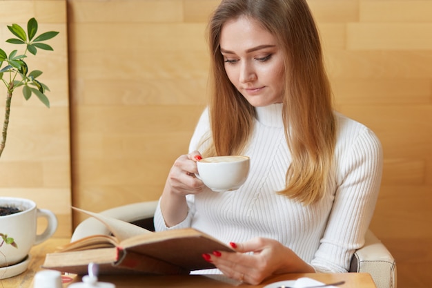La bella giovane donna concentrata legge il libro con una tazza di cappuccino, focalizzata sulla lettura, gode di un interessante romanzo emozionante