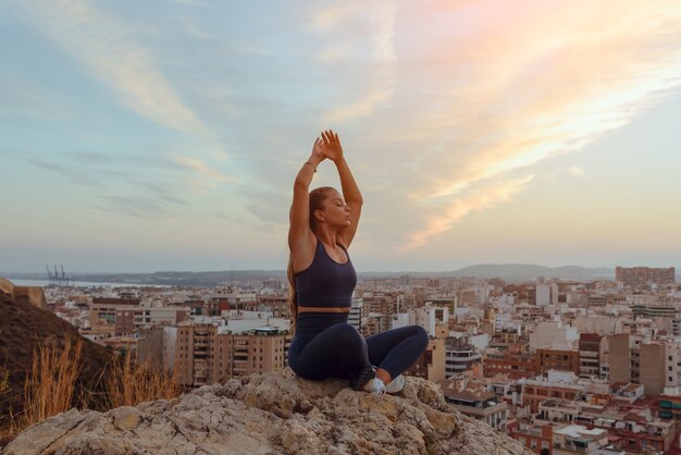 La bella giovane donna fa yoga all'aperto, in cima a un crinale di montagna in città.