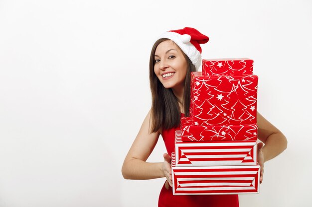 La bella giovane donna felice caucasica con il sorriso affascinante si è vestita in vestito rosso e cappello di Natale che tiene i contenitori di regalo su fondo bianco. Ragazza della Santa con il presente isolato. Concetto di festa di Capodanno 2018