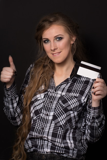 La bella giovane donna in camicia a quadri e con capelli biondi lunghi tiene una carta di credito che sorride e mostra un gesto d'approvazione sfoglia su