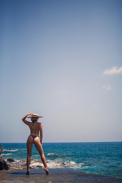 La bella giovane donna in costume da bagno e cappello marroni cammina lungo la spiaggia in riva al mare