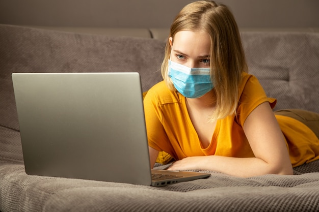 La bella giovane donna in maschera medica blu protettiva ha un lavoro a distanza di studio online sdraiato sul divano. Accogliente ufficio a casa sul divano durante il blocco del coronavirus covid 19. Lavoro freelance utilizzando laptop.