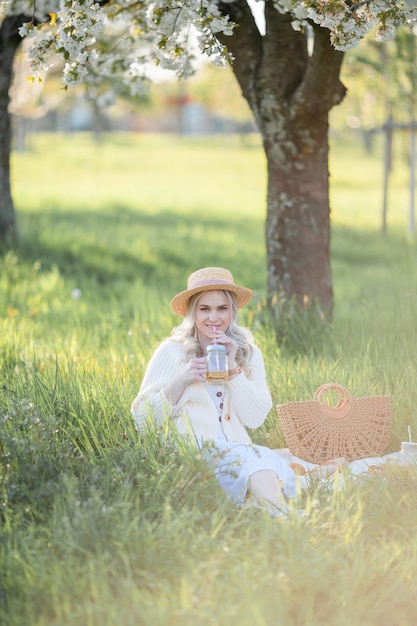 La bella giovane donna in un cappello di vimini sta riposando su un picnic in un giardino fiorito. Fiori bianchi. Primavera. Felicità.
