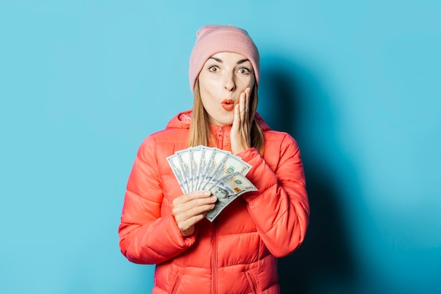 La bella giovane donna in un cappello e una giacca invernale con una faccia sorpresa tiene i soldi