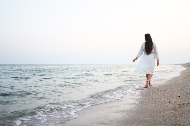 La bella giovane donna in vestito bianco lungo con capelli lunghi e cappello va lungo la spiaggia