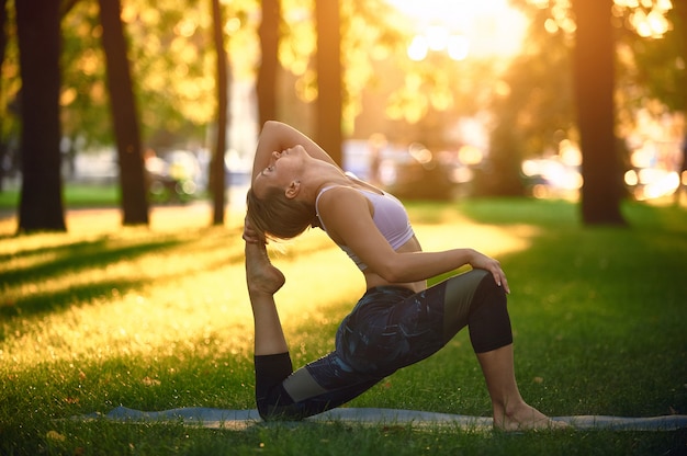 La bella giovane donna pratica il rajakapotasana di posa di re Pigeon di asana di yoga nel parco al tramonto