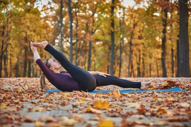 La bella giovane donna pratica l'asana Supta Padangushthasana 1 di yoga sulla piattaforma di legno nel parco di autunno.