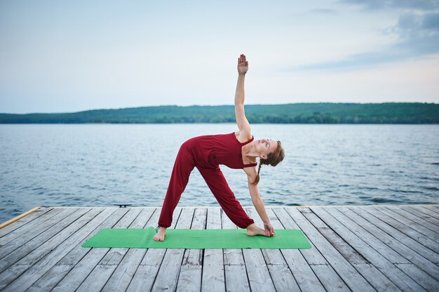 La bella giovane donna pratica lo yoga asana Triangolo posa Trikonasana sul ponte di legno vicino al lago