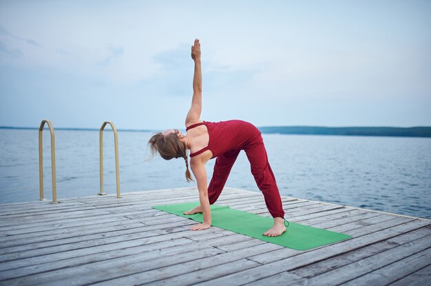 La bella giovane donna pratica lo yoga asana Triangolo posa Trikonasana sul ponte di legno vicino al lago