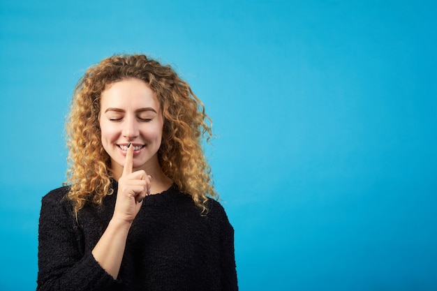 La bella giovane donna riccia rossa positiva con gli occhi chiusi tiene il dito alla bocca, chiede di mantenere un segreto.