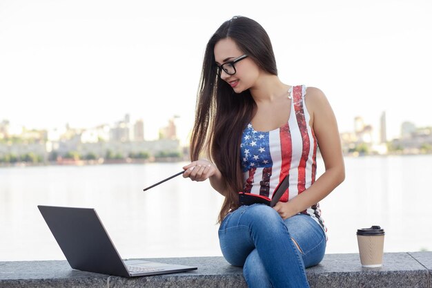 La bella giovane donna si siede nel parco vicino al fiume con il computer portatile.