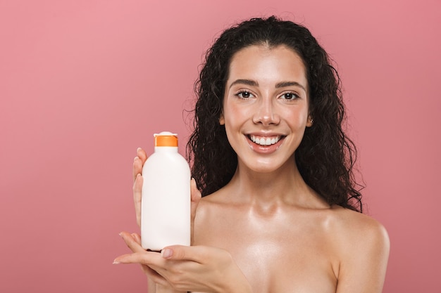 La bella giovane donna sorridente che posa isolata sopra la parete rosa si prende cura della sua pelle che tiene il gel della lozione crema