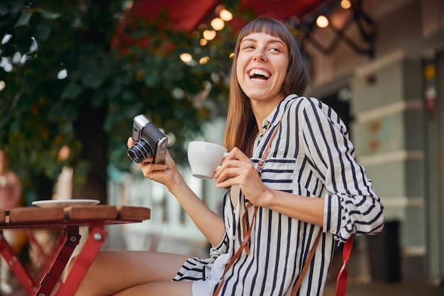 La bella giovane donna sta ridendo mentre tiene la macchina fotografica e la tazza di caffè