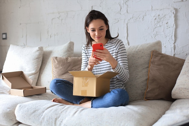La bella giovane donna tiene in mano una scatola di cartone e disimballa lo smartphone seduto sul divano di casa.