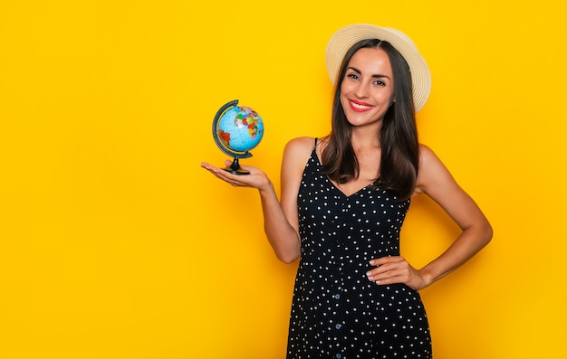 La bella giovane donna turistica castana sorridente in cappello con il globo in mani sta posando su fondo giallo