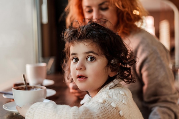 La bella giovane madre dai capelli rossi con la figlia carina dai capelli ricci è seduta in un caffè accogliente e beve la calda festa della mamma al caldo della scuola