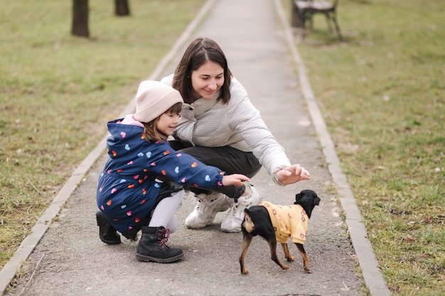 La bella giovane mamma cammina con la figlia e la famiglia felice del piccolo cane che cammina all'aperto con l'animale domestico durante