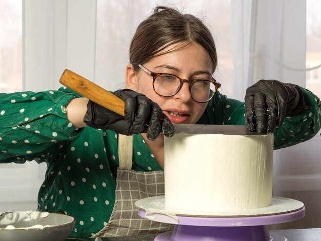 La bella giovane pasticcera fa una torta con crema bianca usando una spatola da cucina