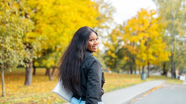 La bella giovane ragazza afroamericana con un sorriso in vestiti alla moda cammina in un parco di autunno con fogliame giallo stupefacente
