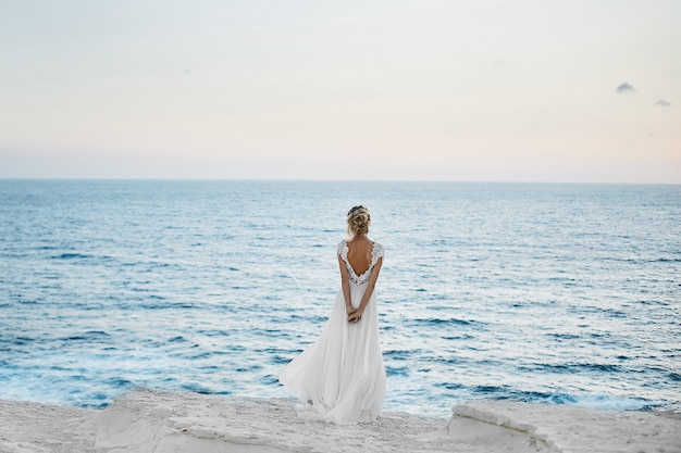 La bella giovane ragazza di modello bionda in vestito bianco sta indietro e guarda il mare