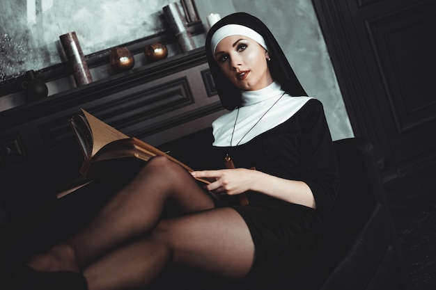 La bella giovane suora in vestito nero di religione tiene la Bibbia e posa sulla macchina fotografica con un grande libro su uno sfondo nero. Avvicinamento. concetto di religione.
