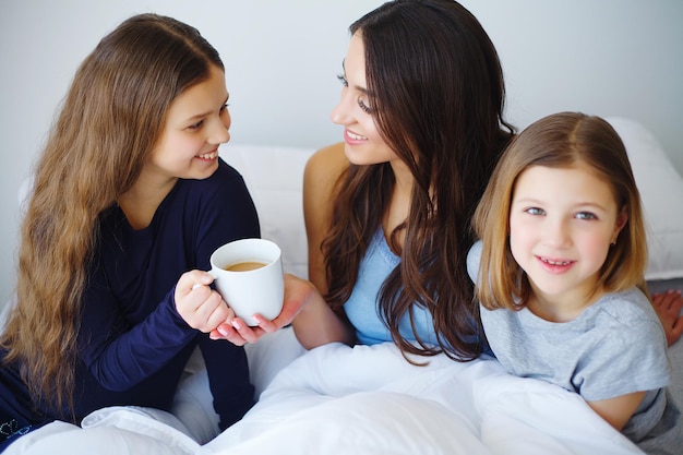 La bella mamma e le figlie si abbracciano, guardano la macchina fotografica e sorridono mentre sono sedute sul letto a casa.